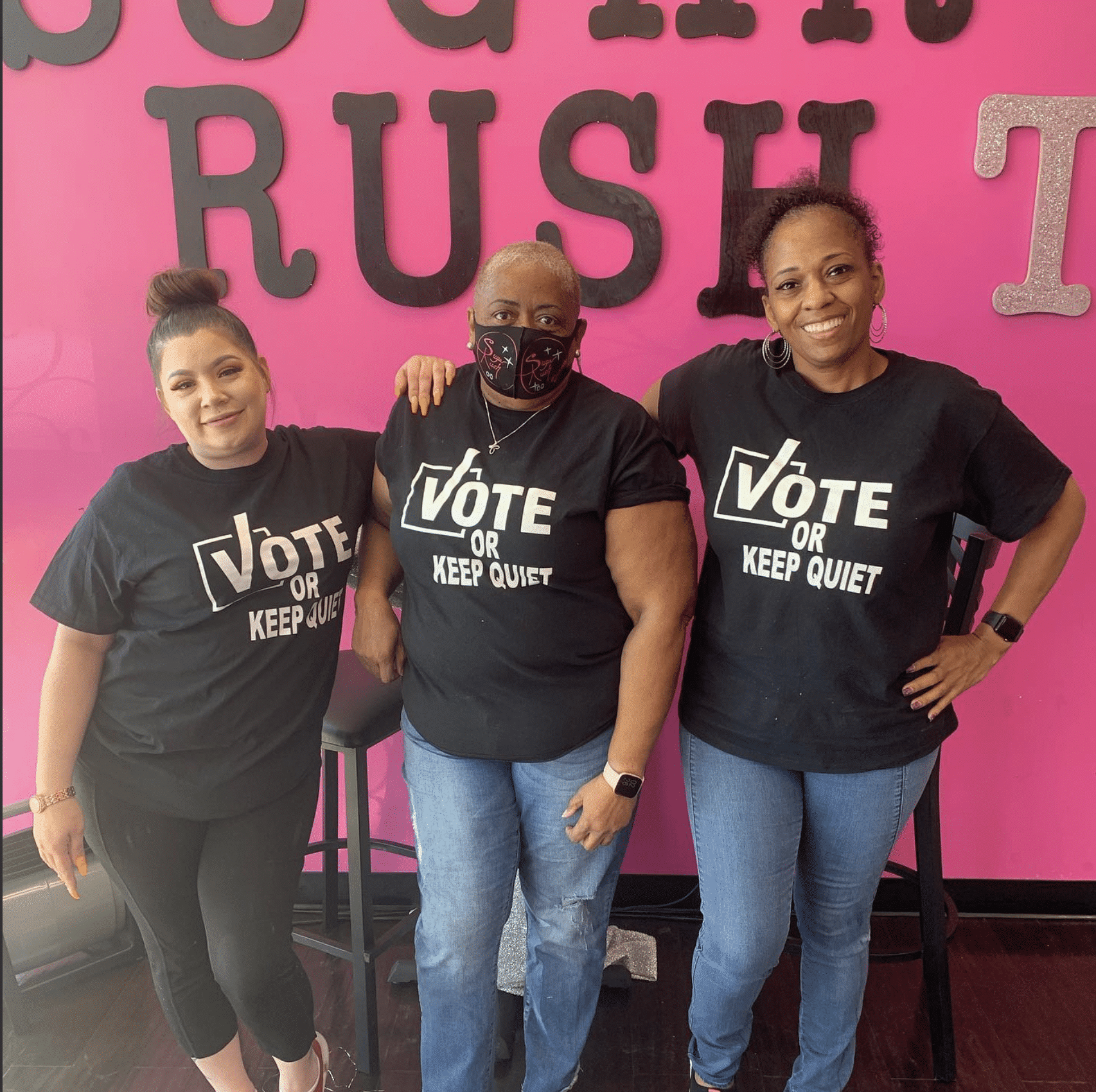 Three women posing in front of a sign at Sugar Rush Too OST bakery. They sell some of the best tea cakes and they are a black owned business. 