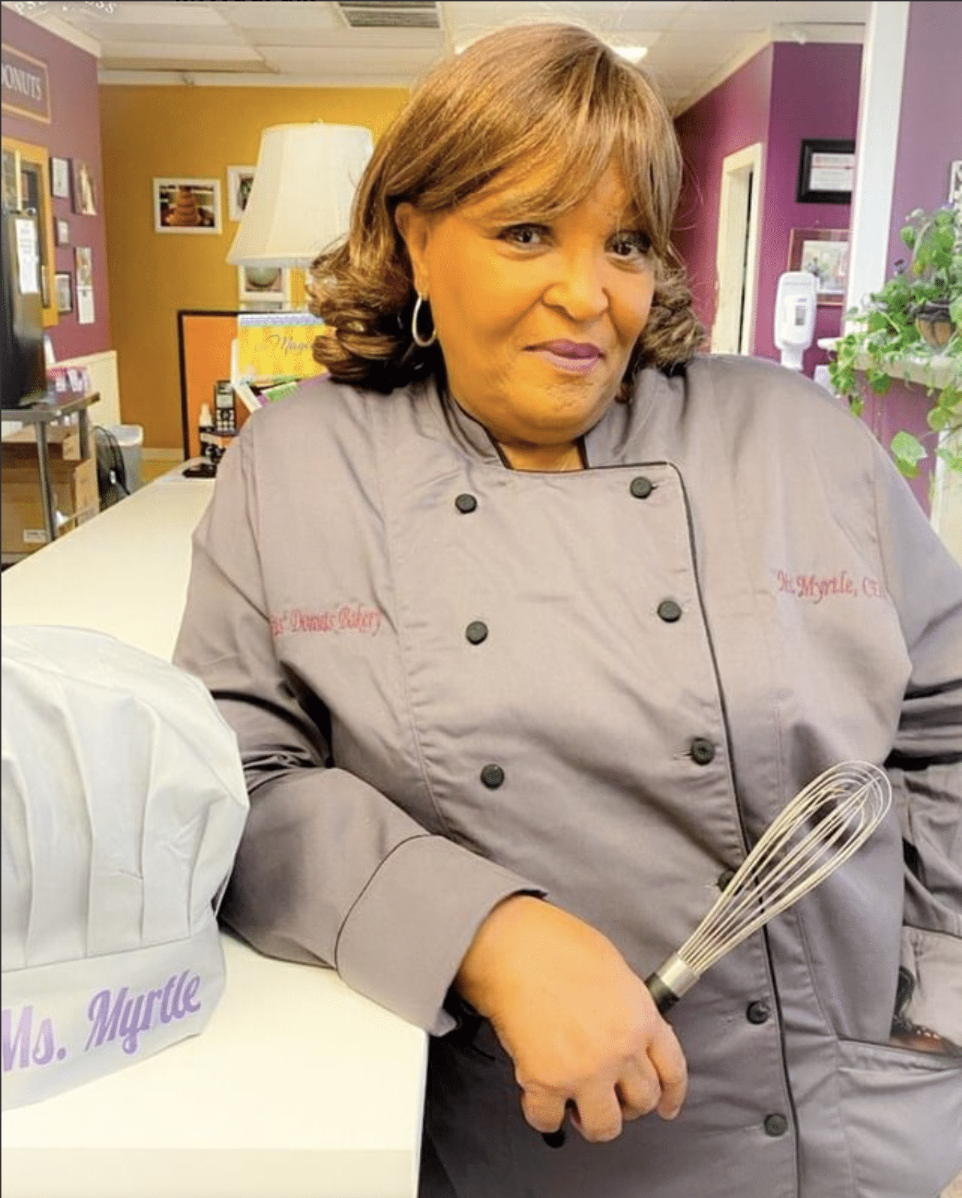 Ms. Myrtle of Ms. Myrtle's Bakery Shoppe, formerly Not Jus Donuts, leaning on a counter while wearing a chef's coat and holding a whisk. A baker's hat with her name embroidered on it sits beside her. She makes some of the best tea cakes in Houston. 