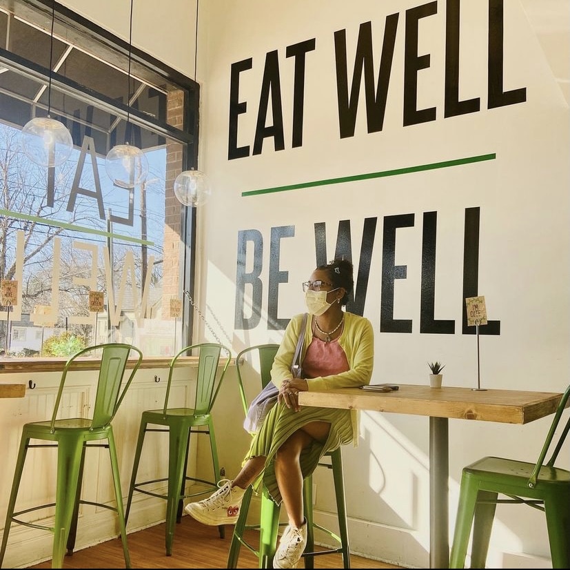 A woman in a mask sits alone in Local Green Atlanta, against a backdrop that says Eat Well, Be Well.