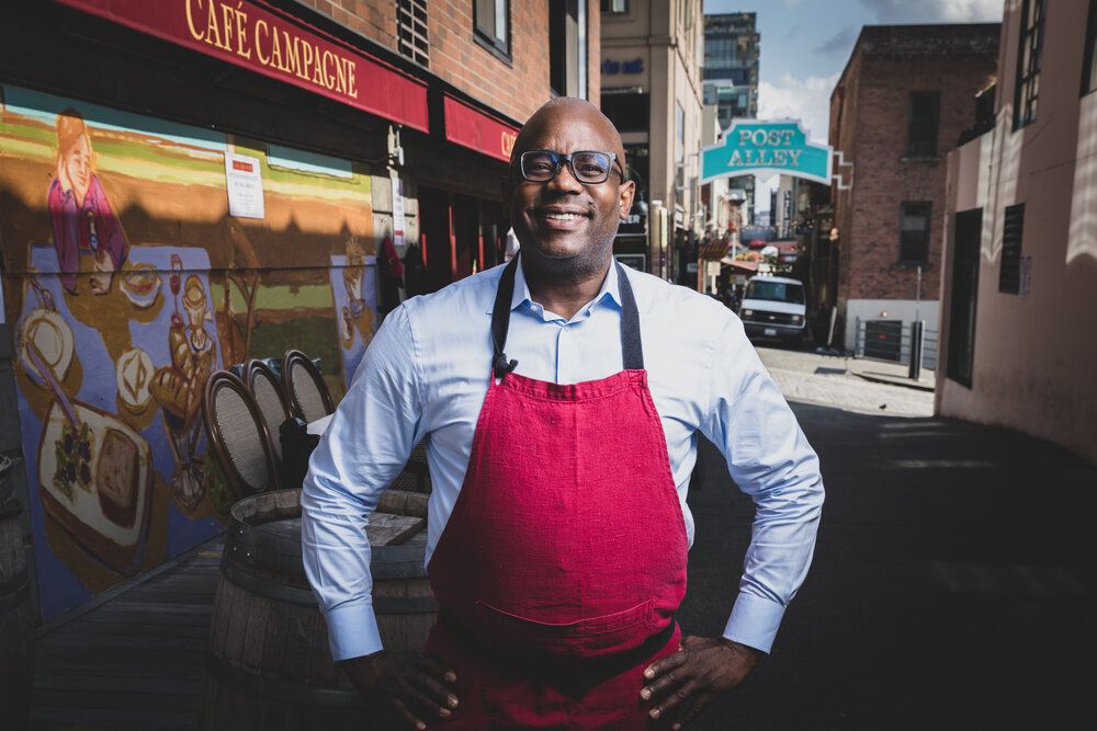 Chef Daisley Gordon smiles wide in a red chef's apron in front of Cafe Campagne in Seattle. This Black-owned breakfast restaurant is serving up some of the best French-inspired omlettes and French Toast in Seattle.