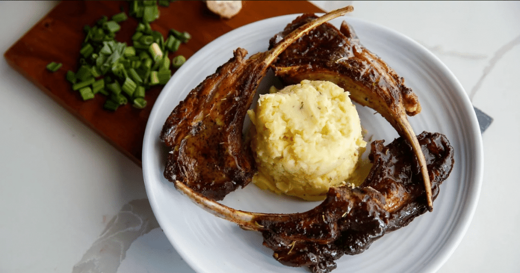 Three succulent lamb chops encircle a mound of mashed potatoes, which sits beside a chopping board with various ingredients. This elevated soul food is from Esther's Cajun Cafe, one of the best new restaurants in Houston in 2022. This is also a new Black-owned restaurant in Houston.